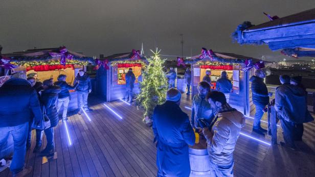 Menschen besuchen einen Weihnachtsmarkt auf einer Dachterrasse bei Nacht.