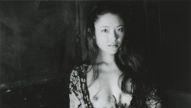 Eine Frau mit offenem Haar und einer floralen Jacke vor dunklem Hintergrund.