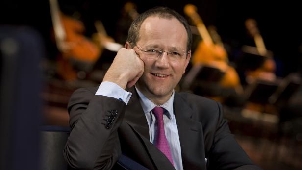 Ein lächelnder Mann mit Brille und Anzug sitzt vor einem Orchester.