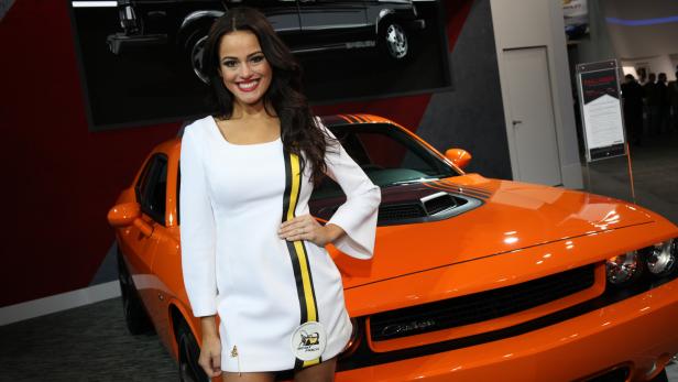 Eine Frau posiert vor einem orangefarbenen Dodge Challenger auf einer Automobilmesse.