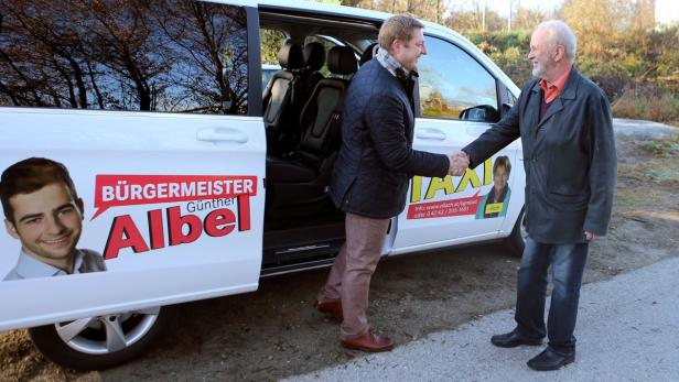 Zwei Männer schütteln sich vor einem Taxi mit Wahlwerbung für Bürgermeister Günther Albel die Hände.