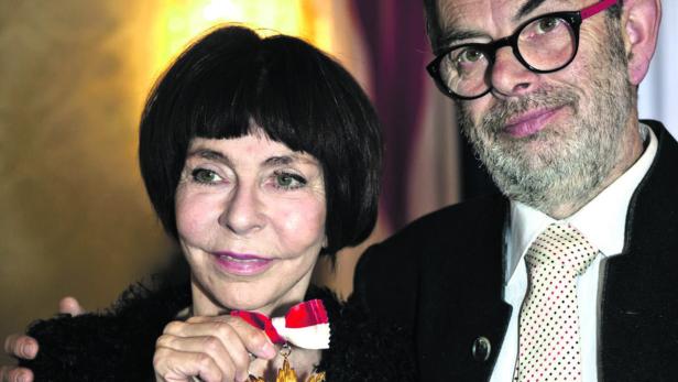 Agnès Varda hält eine Auszeichnung, während ein Mann im Hintergrund steht.