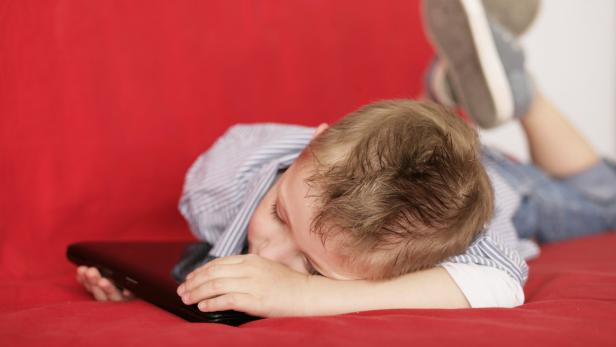 Ein Junge liegt erschöpft mit dem Gesicht auf einem Laptop auf einem roten Sofa.