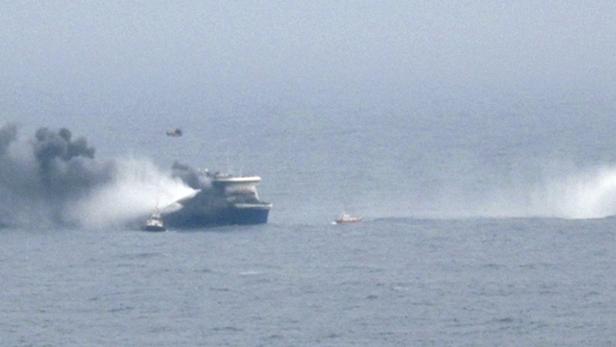 Ein Schiff auf dem Meer wird mit Wasserwerfern gelöscht, während ein Hubschrauber darüber fliegt.