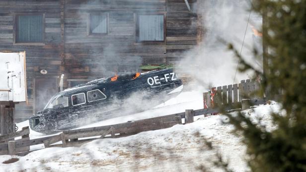 Ein abgestürztes Flugzeug mit der Kennung OE-FZ liegt rauchend vor einem Holzhaus im Schnee.