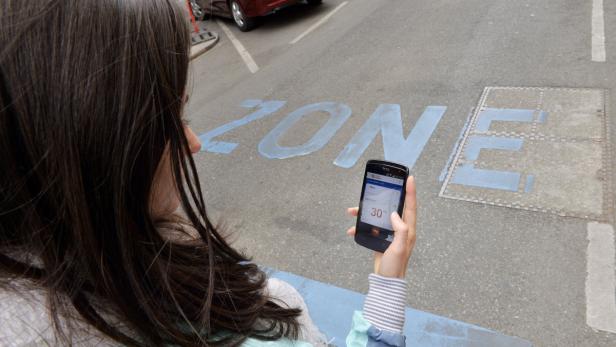Eine Frau betrachtet ein Smartphone vor einer blau markierten Zone auf der Straße.