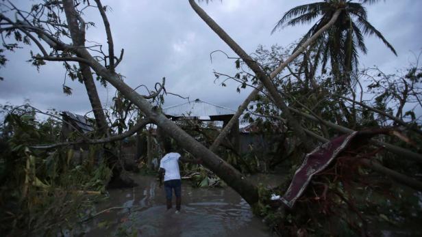 Ein Mann watet durch das Wasser inmitten von umgestürzten Bäumen und beschädigten Häusern.