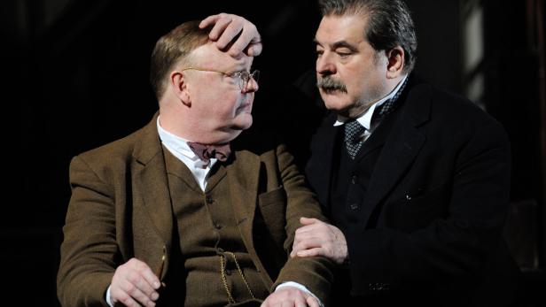 Zwei Männer in Anzügen interagieren auf einer Bühne.