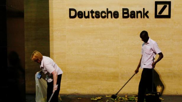 Zwei Reinigungskräfte fegen Laub vor einer Filiale der Deutschen Bank.