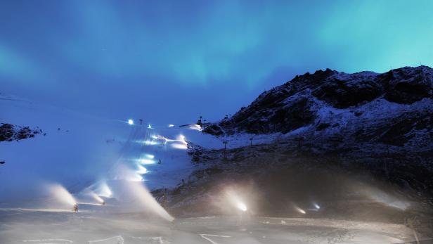 Schneekanonen erzeugen Kunstschnee auf einer Skipiste unter einem Nachthimmel mit Nordlichtern.