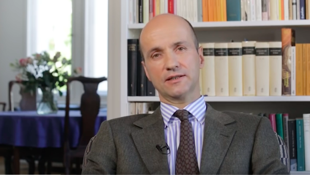 Ein Mann mit Anzug und Krawatte sitzt vor einem Bücherregal.