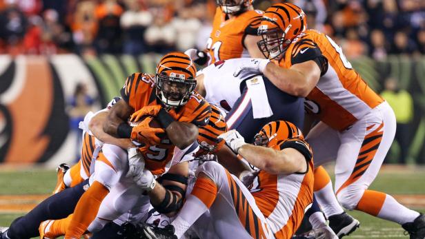 Ein Footballspieler in orangefarbener Uniform wird von mehreren Gegenspielern zu Fall gebracht.