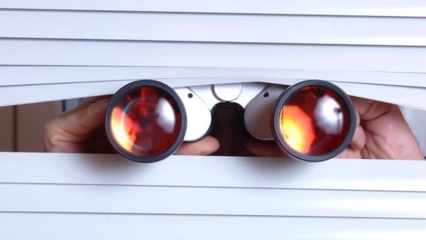 Jemand späht mit einem Fernglas durch die Jalousien.