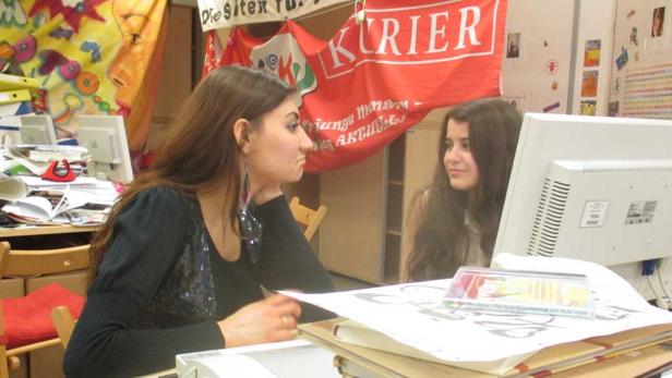 Zwei junge Frauen sitzen an einem Schreibtisch mit einem „Kurier“-Banner im Hintergrund.