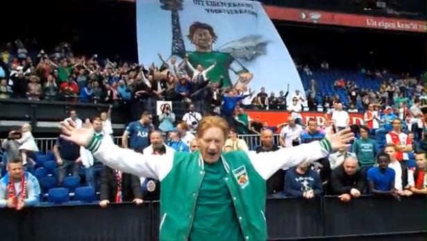 Ein Mann in einer grünen Jacke steht vor einer jubelnden Menschenmenge in einem Stadion.