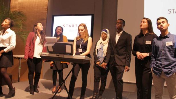 Eine diverse Gruppe junger Leute singt gemeinsam vor einem Keyboard auf einer Bühne.