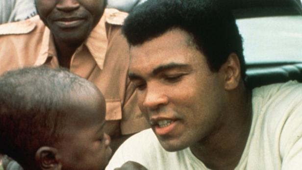 Muhammad Ali interagiert mit einem Kind, während eine weitere Person im Hintergrund steht.