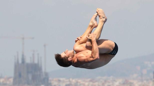Ein männlicher Turmspringer in der Luft, im Hintergrund die Sagrada Familia in Barcelona.