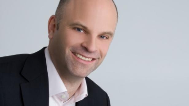 Ein lächelnder Mann mit Anzug vor einem hellen Hintergrund.