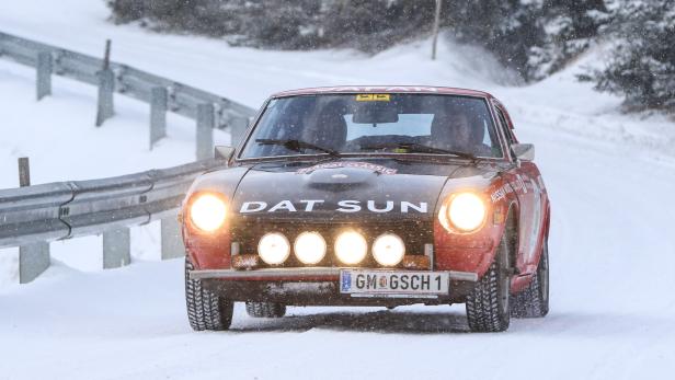 Ein roter Datsun fährt mit eingeschalteten Scheinwerfern auf einer schneebedeckten Straße.