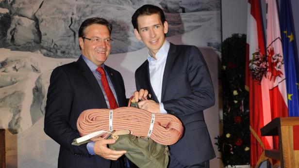 Zwei Männer im Anzug halten eine Kletterausrüstung vor einer Bergkulisse.