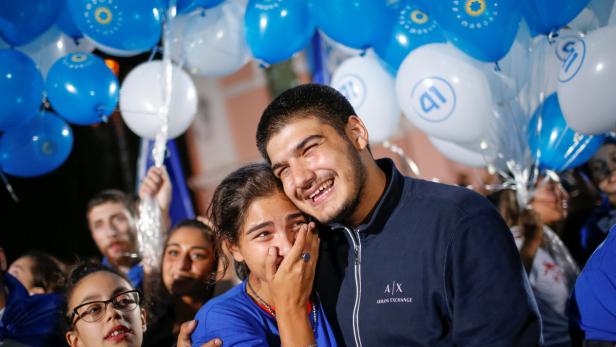 Ein lächelnder Mann umarmt ein weinendes Mädchen vor einem Hintergrund aus blauen und weißen Luftballons.