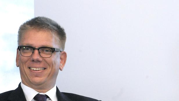 Ein lächelnder Mann mit Brille und Anzug vor dem OMV-Logo.