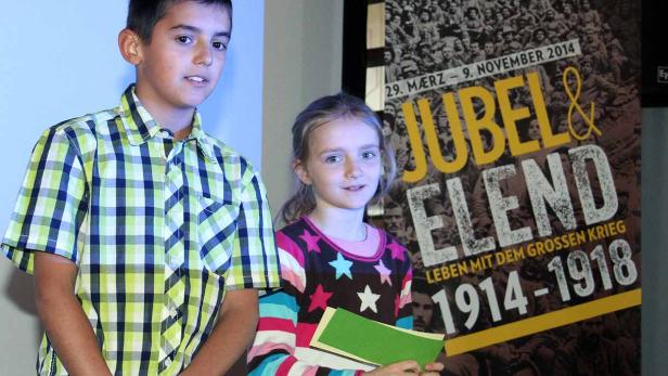 Ein Junge und ein Mädchen stehen vor einem Plakat der Ausstellung „Jubel & Elend 1914-1918“.