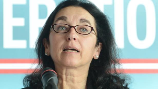 Eine Frau mit Brille spricht in ein Mikrofon vor einem hellblauen Hintergrund.