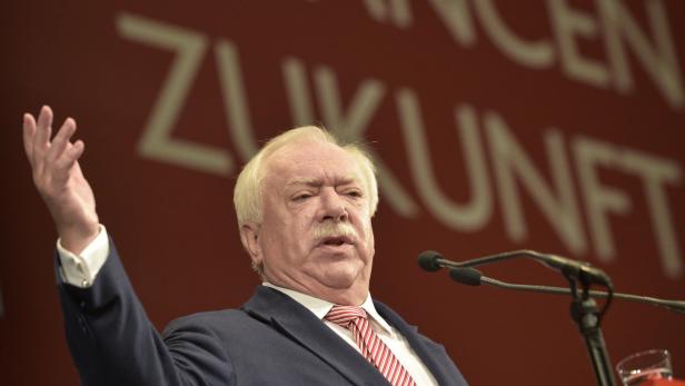 Ein Mann mit Schnurrbart gestikuliert vor einem roten Hintergrund mit dem Wort „Zukunft“.