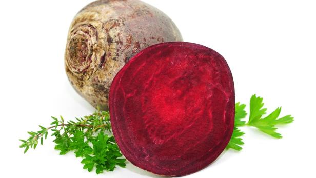 Eine ganze und eine halbe rote Bete liegen auf einem weißen Untergrund, garniert mit Kräutern.
