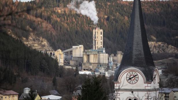 Eine Fabrik raucht vor einem bewaldeten Hügel, im Vordergrund ein Kirchturm.