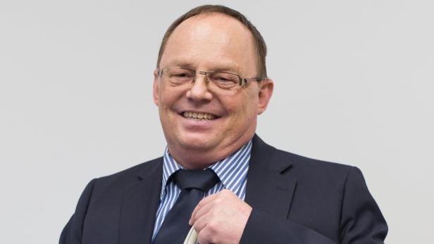 Ein lächelnder Mann mit Brille, Anzug und Krawatte vor einem hellen Hintergrund.