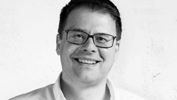 Ein lächelnder Mann mit Brille vor einer weißen Wand.