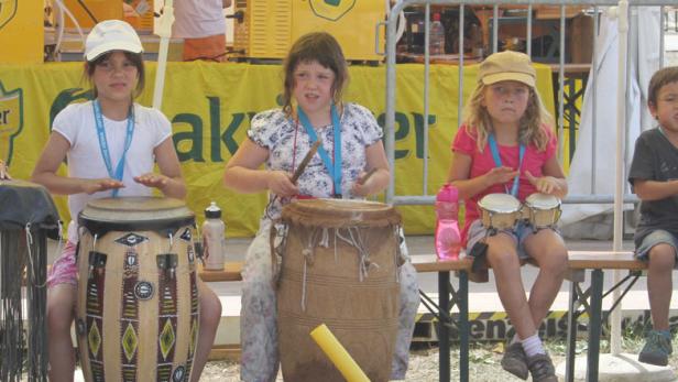 Eine Gruppe von Kindern sitzt auf einer Bank und spielt auf verschiedenen Trommeln.