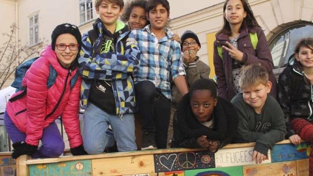 Eine Gruppe von Kindern posiert vor einem bemalten Holzzaun mit der Aufschrift „Refugees Welcome“.