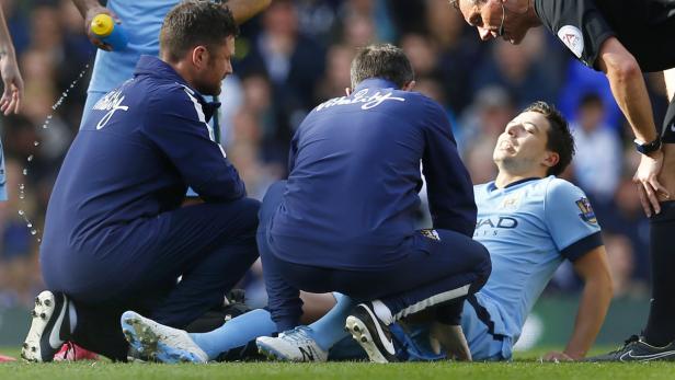 Ein Fußballspieler liegt verletzt auf dem Rasen, während er von Sanitätern und einem Schiedsrichter betreut wird.