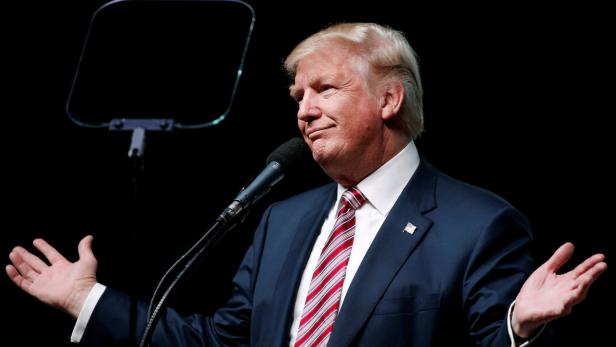 Donald Trump gestikuliert während einer Rede vor einem dunklen Hintergrund.