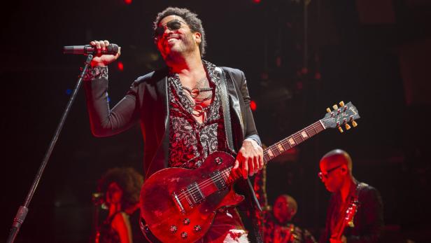 Lenny Kravitz tritt mit Sonnenbrille, Gitarre und Mikrofon auf einer Bühne auf.