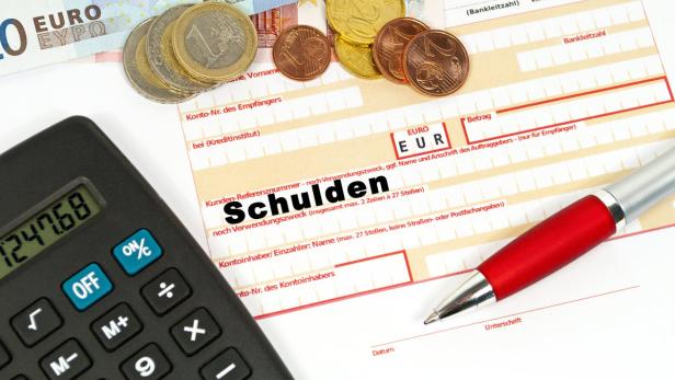 Ein Überweisungsformular mit dem Wort „Schulden“, Euro-Münzen, einem Schein, einem Taschenrechner und einem Stift.