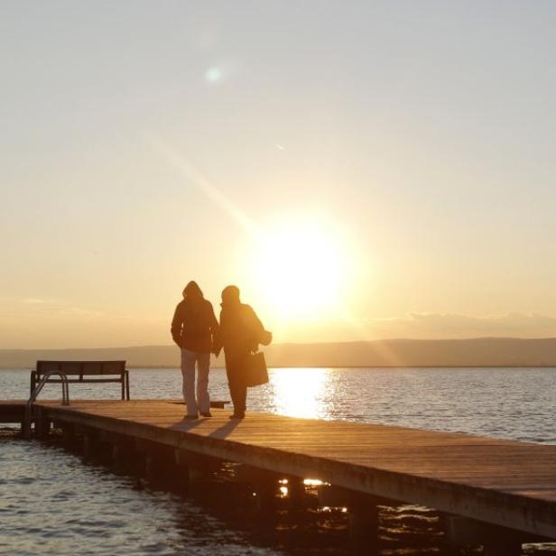 Zwei Personen gehen bei Sonnenuntergang auf einem Steg am Wasser entlang.
