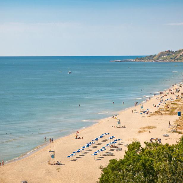 Ein belebter Sandstrand mit vielen Menschen und blau-weißen Sonnenschirmen an einem sonnigen Tag.