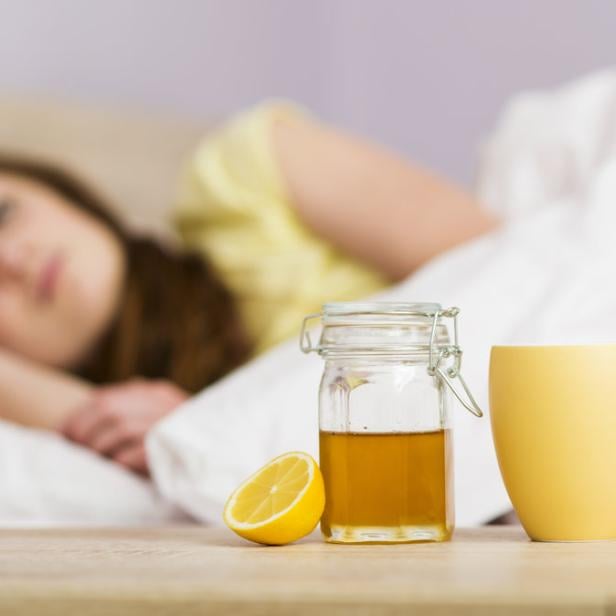 Eine kranke Frau liegt im Bett, neben ihr stehen Honig, Zitrone und eine Tasse.