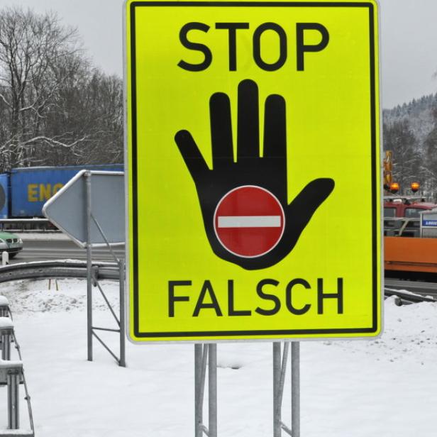 Ein gelbes Schild mit der Aufschrift „STOP FALSCH“ und einer Hand mit einem roten Kreis darauf steht an einer winterlichen Straße.
