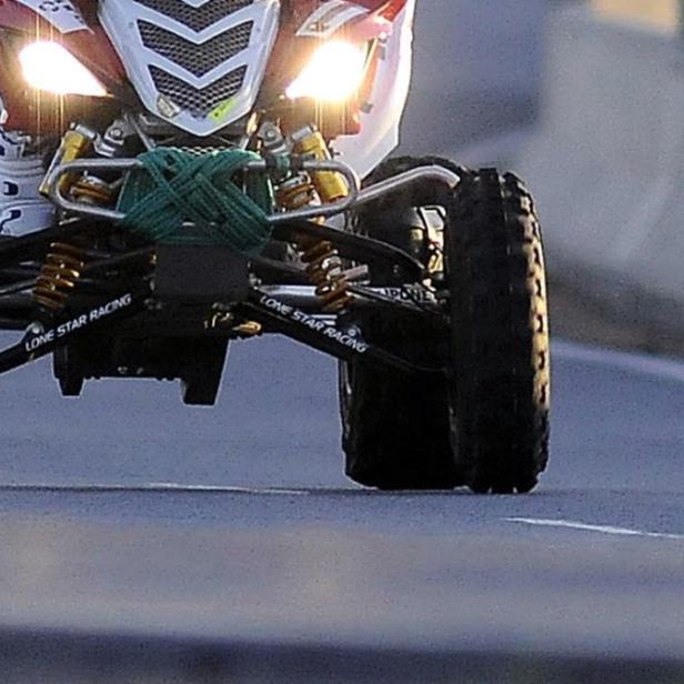 Ein Quad mit eingeschalteten Scheinwerfern fährt auf einer asphaltierten Straße.
