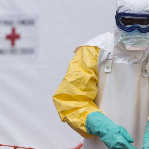 Eine Person in Schutzkleidung und Schutzbrille vor einem Zelt mit einem roten Kreuz.