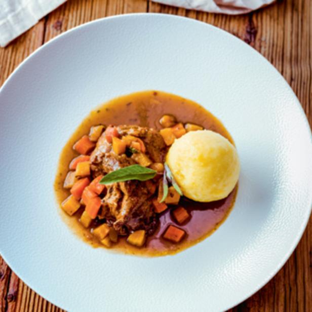 Ein Teller mit Ossobuco, Gemüsewürfeln und einem gelben Knödel auf einem Holztisch.
