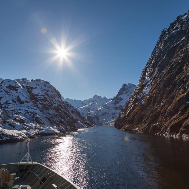 Blick vom Bug eines Schiffes auf einen Fjord mit schneebedeckten Bergen unter strahlender Sonne.