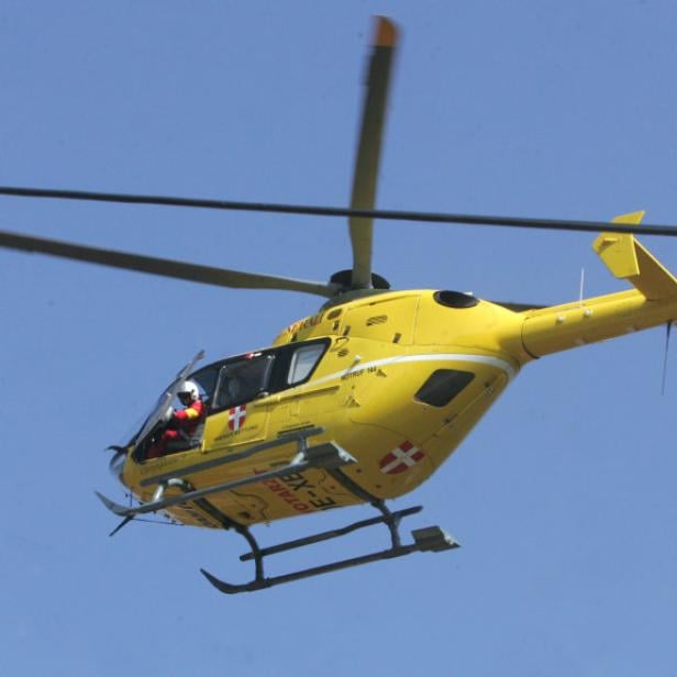 Ein gelber Rettungshubschrauber mit österreichischer Flagge am Heck fliegt vor blauem Himmel.