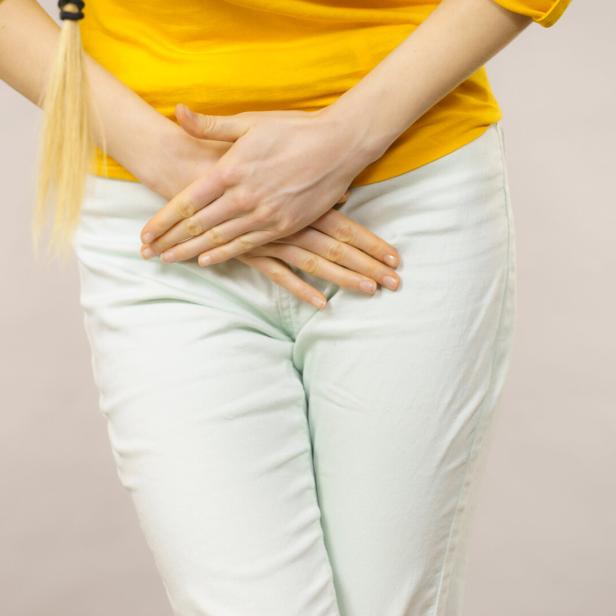 Eine Person hält sich mit beiden Händen den Unterbauch und trägt ein gelbes Oberteil sowie eine helle Hose.